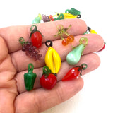 purple glass grapes, red glass berries, red glass cherries, yellow glass bananas, green glass pear, green glass bell pepper, red glass creole tomato, and red glass strawberry in a hand on a white background