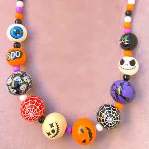 painted wooden beads with a Halloween theme. Beads include an eyeball, the word "boo", a scream face, spiderweb, jack-o-lantern faces, a witch on a broom and a black cat face. Beads are paired with purple, orange, black and white beads