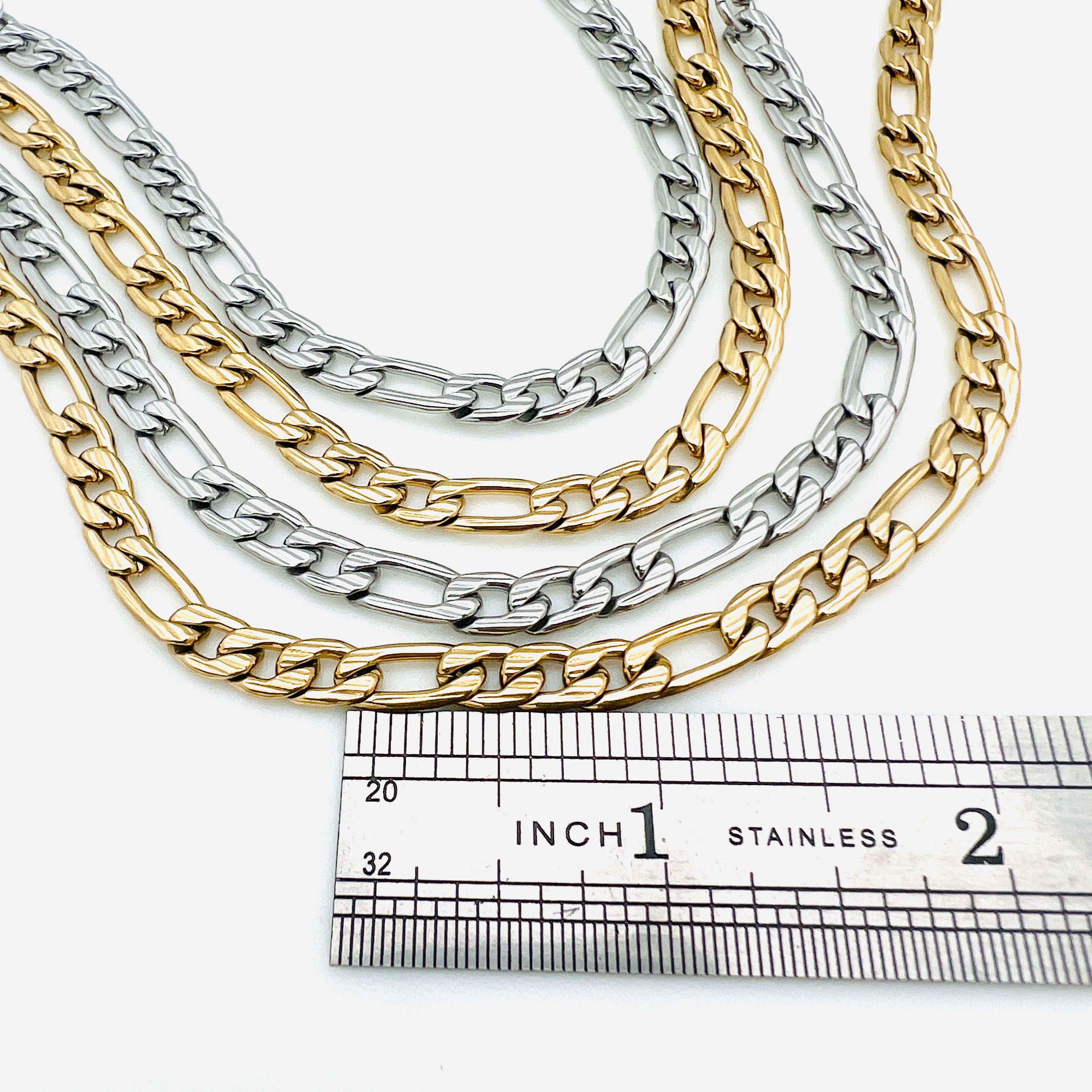 two silver tone and two golden textured Figaro link chains displayed above a stainless steel ruler on a white background.
