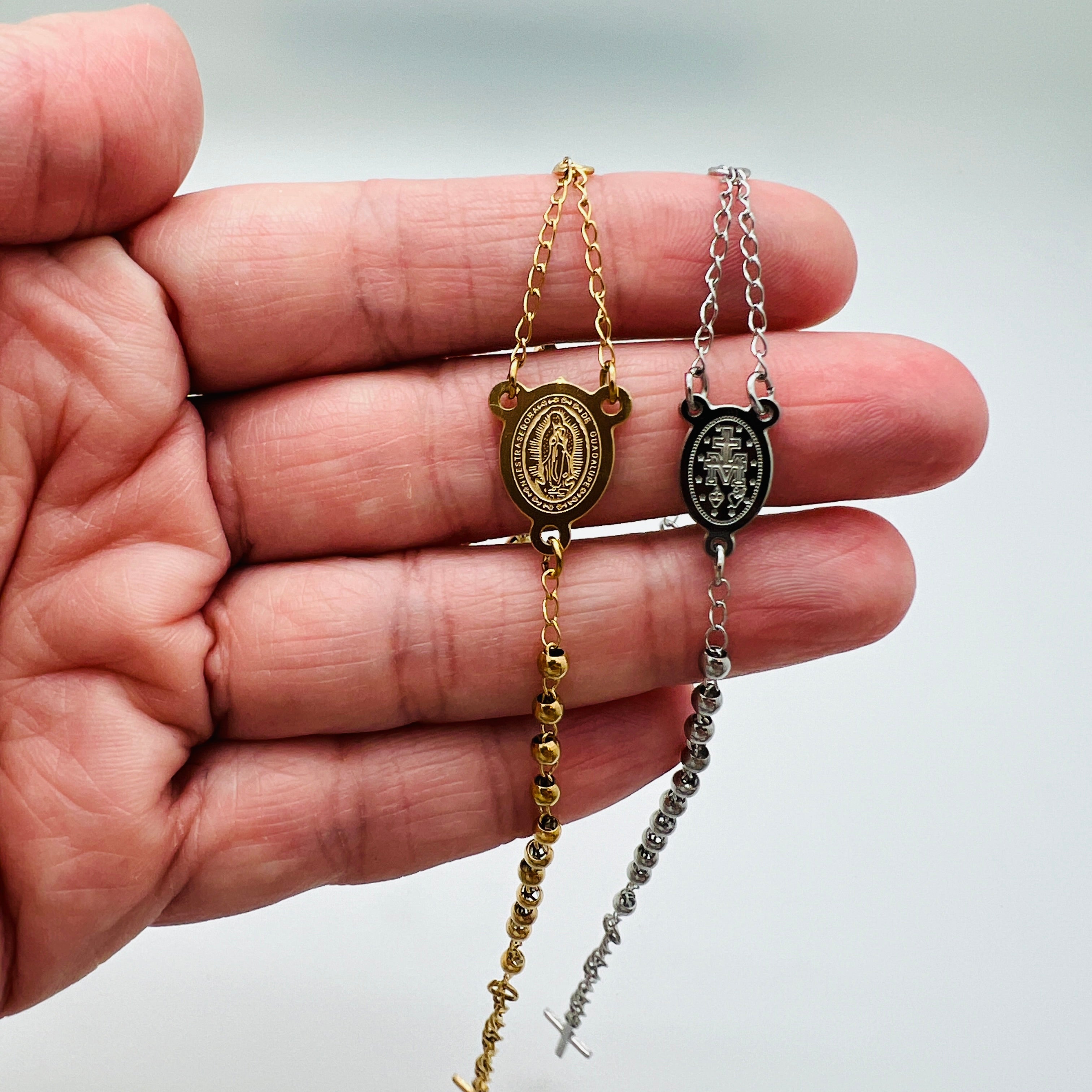 golden and silver tone rosary bracelets with Virgin Mary connector piece a ball link chain draped over hand with white background,. 
