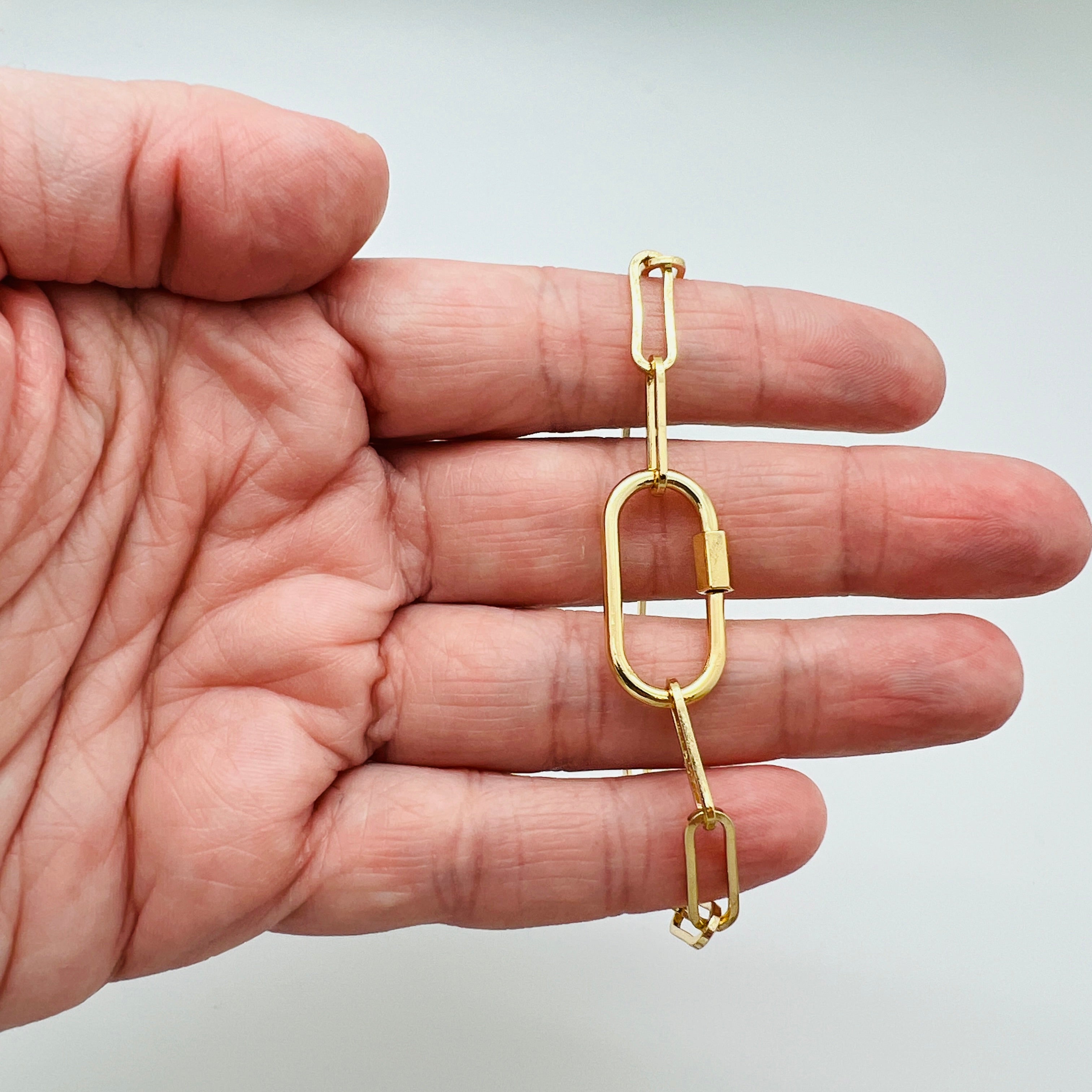 golden paperclip chain bracelet with carabiner clasp draped over a hand with white background. 