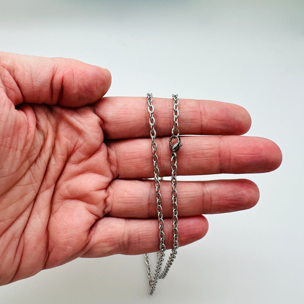 silver tone flat cable chain necklace with lobster claw clasp draped over a hand over white background. 