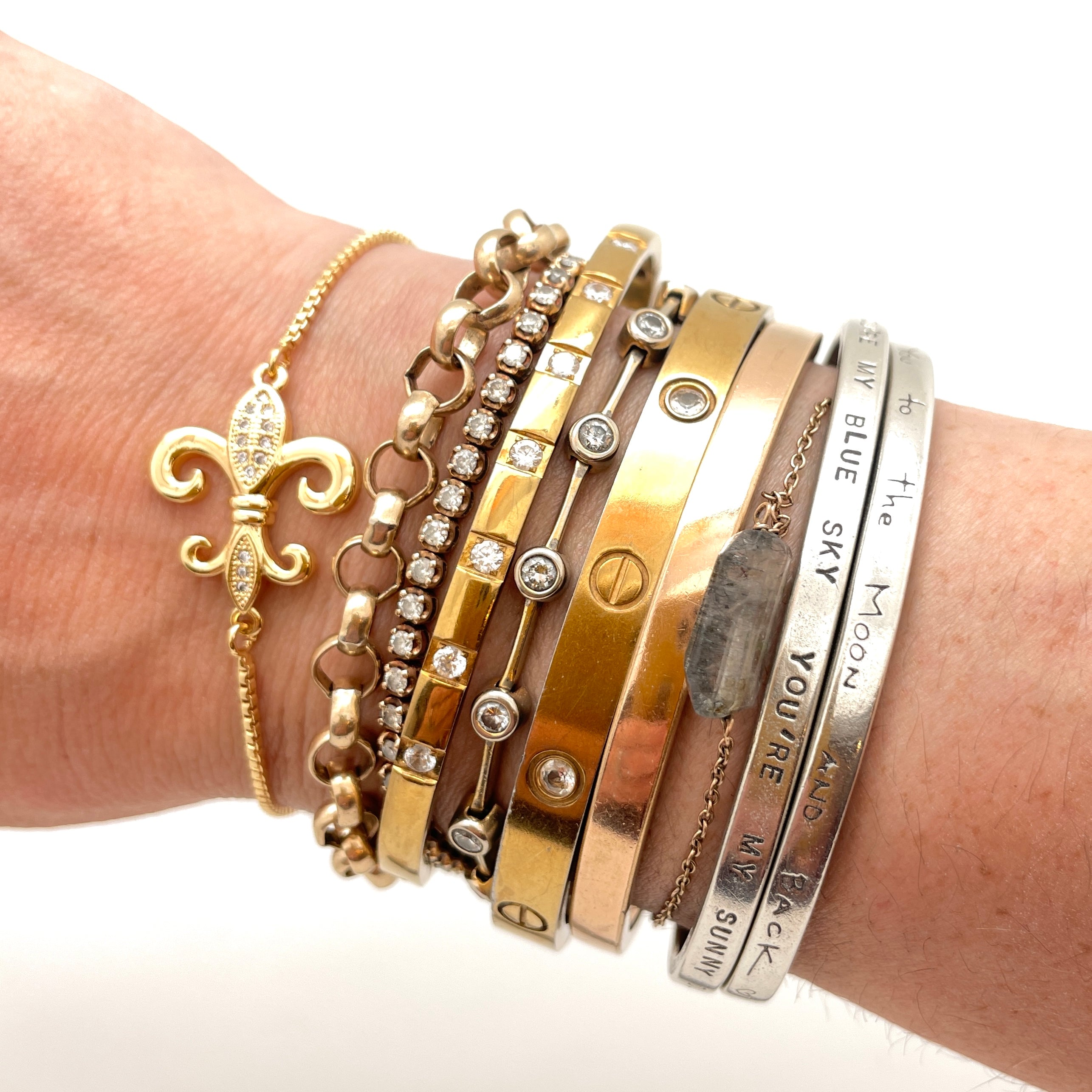 golden fleur de lis bracelet stacked amongst nine other bracelets of varying styles and metals on a wrist with a white background. 