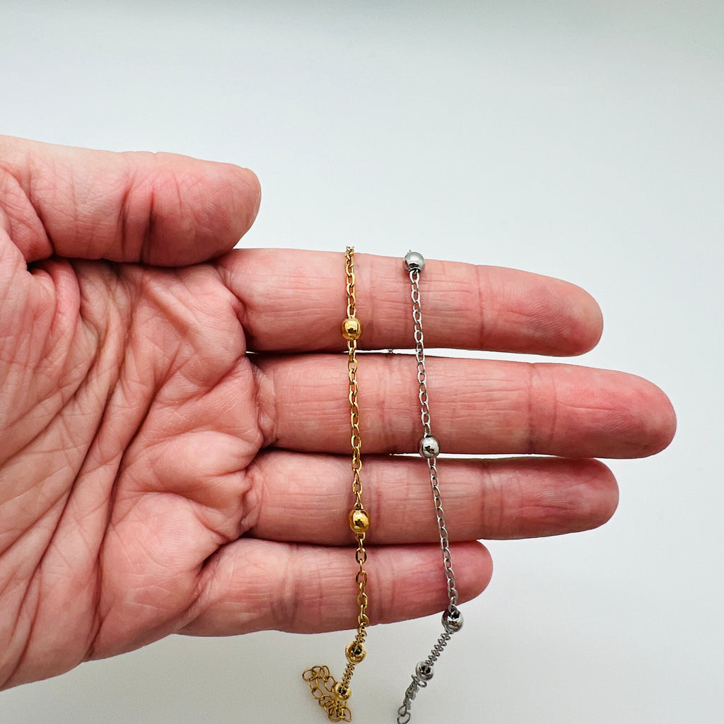 golden and silver tone satellite chain anklets draped over hand with white background. 