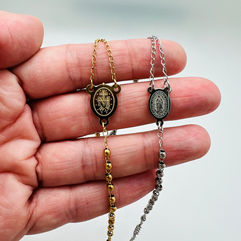 golden and silver tone rosary bracelets with Virgin Mary connector piece a ball link chain draped over hand with white background,. 