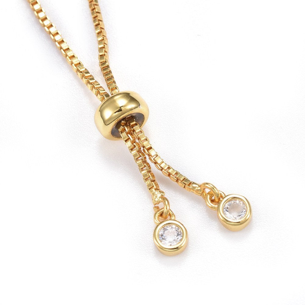 close-up of adjustable golden box chain with slider bead and two bezel set clear stone charms over white background. 