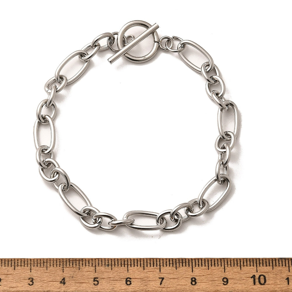silver Figaro chain bracelet with toggle clasp displayed above a wooden imperial ruler on white background. 