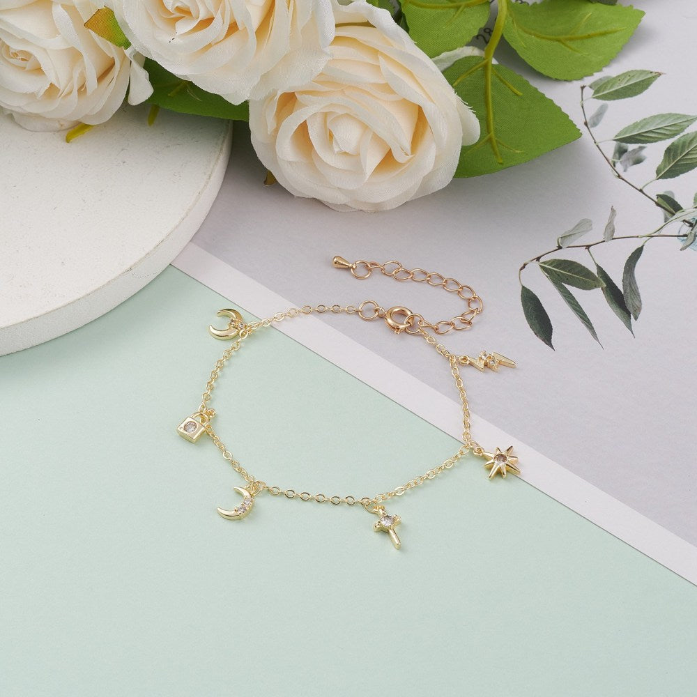 golden and clear stone charm bracelet with spring ring clasp and extender chain including a lightening bolt, star, cross, moon, padlock and crescent charms over grey, white and mint green surface with white flowers and green leaves peeking into the frame. 