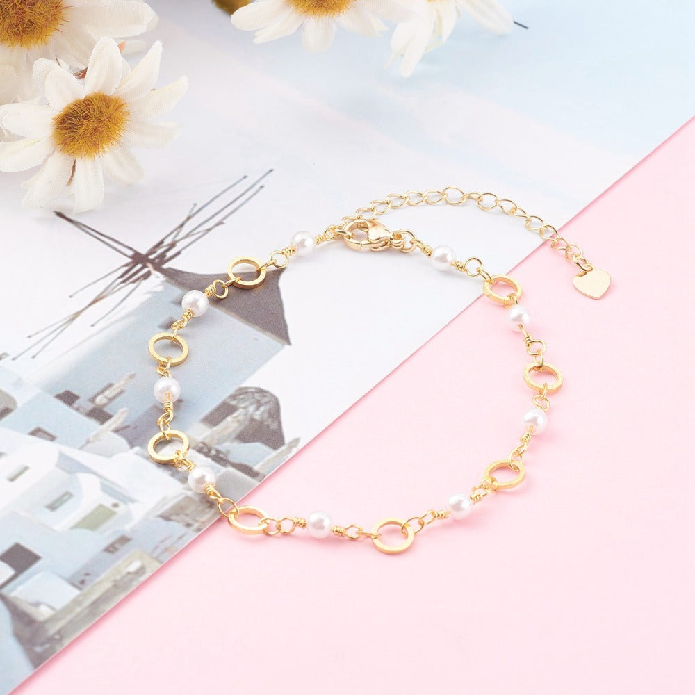 golden chain bracelet alternating ring and white faux pearl links with lobster claw clasp and extender chain over light pink surface with a photo of white buildings and sky with daisies peeking in from the top left corner. 
