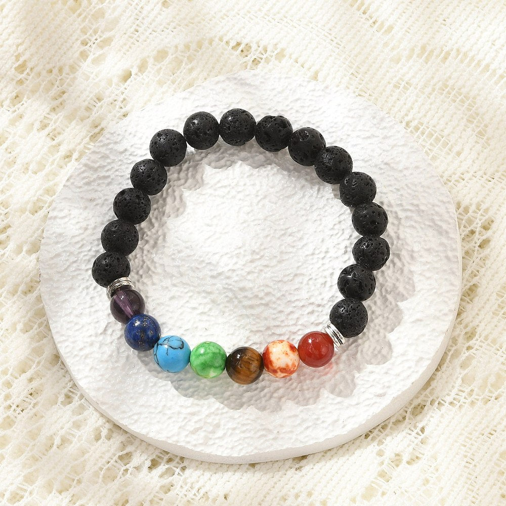 black lava rock stretch chakra bracelet featuring seven round chakra stone beads between silver double line spacer beads on a white textured coasted over off white lace surface.