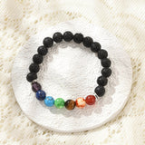 black lava rock stretch chakra bracelet featuring seven round chakra stone beads between silver double line spacer beads on a white textured coasted over off white lace surface.