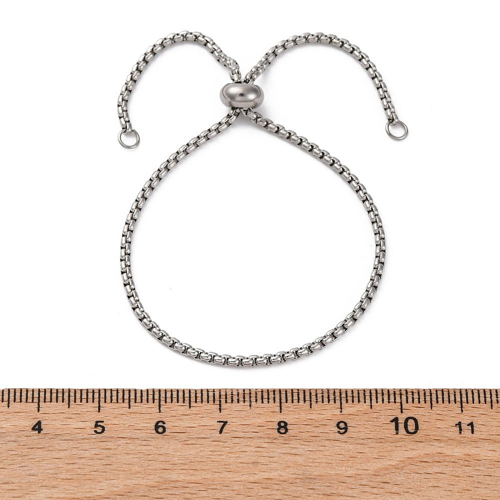 silver tone bolo bracelet with stopper bead and jump rings on ends displayed over wooden ruler with white background .