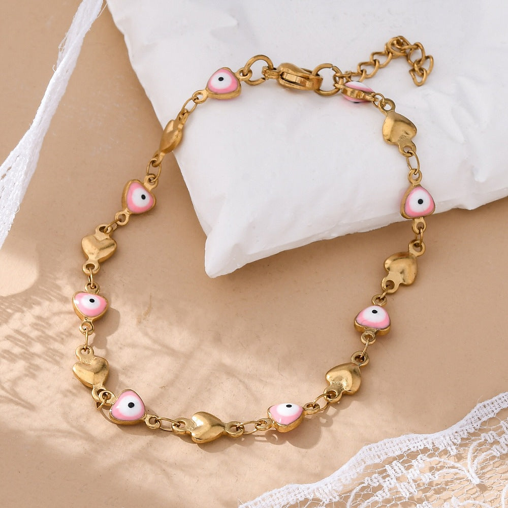 golden heart link bracelet featuring a pink evil eye on every other link plus a lobster claw clasp and extender chain draped over a white pillow onto a tan surface. 