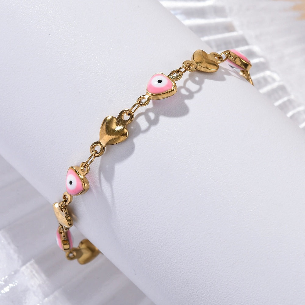 golden heart link bracelet featuring a pink evil eye on every other link wrapped around white cylinder form over shiny white surface. 