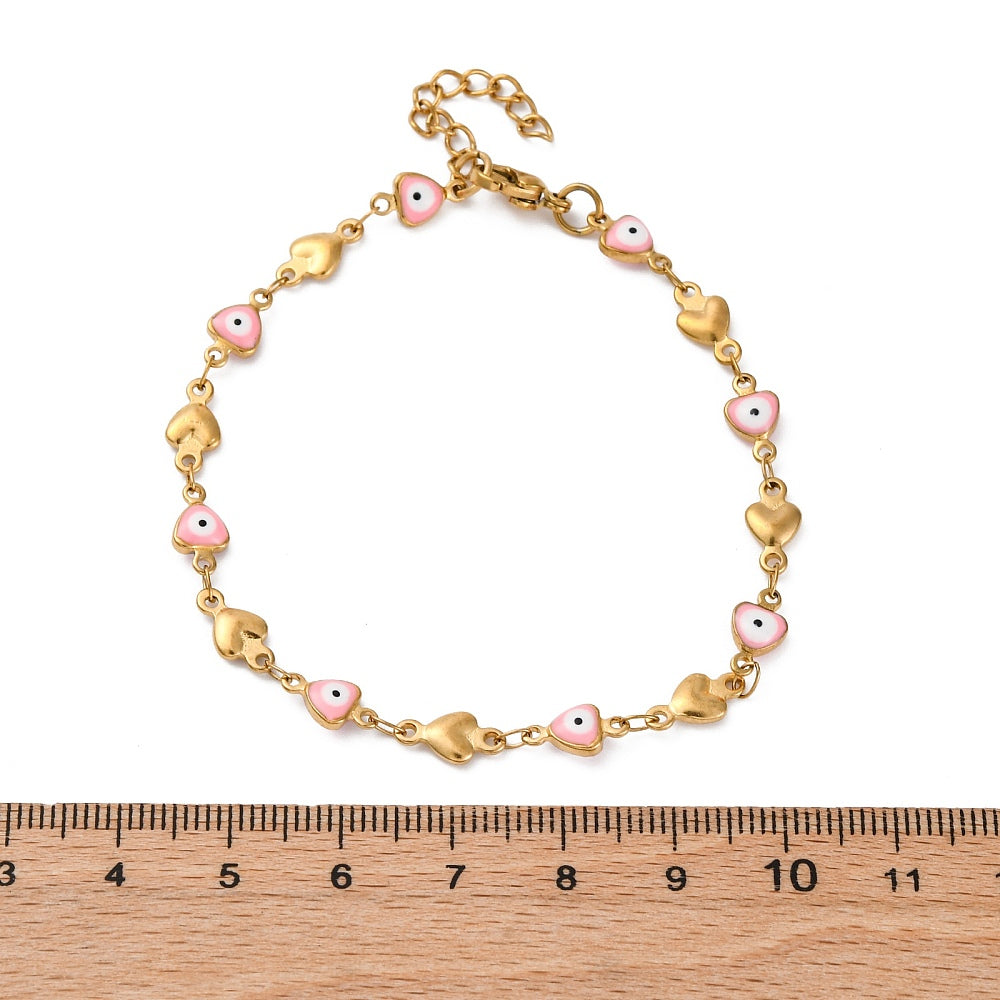 golden heart link bracelet featuring a pink evil eye on every other link plus a lobster claw clasp and extender chain above wooden ruler with white background. 