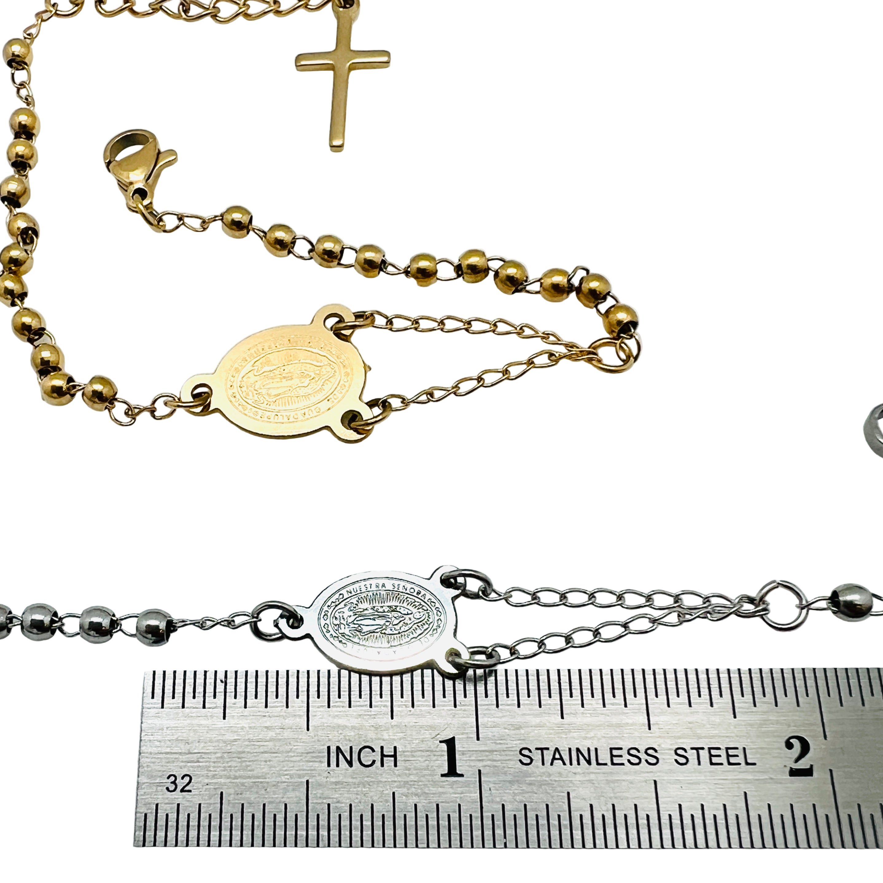 golden and silver tone rosary bracelets with Virgin Mary connector piece a ball link chain with lobster claw claps and extender chain with a dangling cross charm displayed above a ruler with white background,. 
