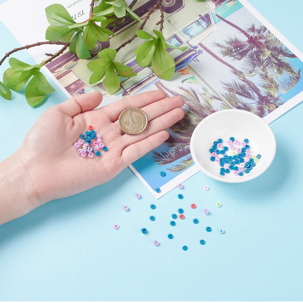 a hand with some examples of loose beads from the kit in it with a dollar coin and next to the hand is a small white bowl and some more loose beads on a light blue surface with a green leafy branch peeking into the frame. 