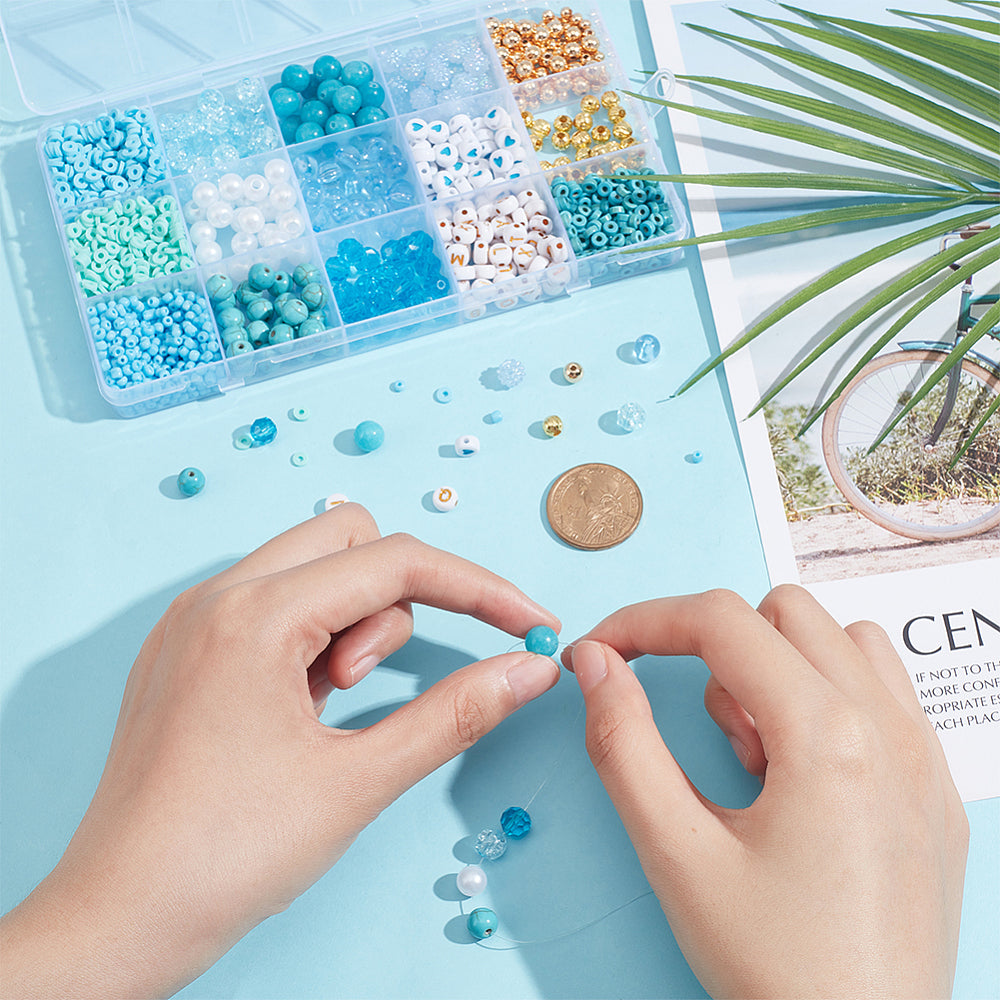 the plastic bead box filled with a variety of blue and gold beads along with two hands creating a design with a dollar coin between the box and hands and some loose beads around all over a light blue surface. 