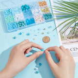 the plastic bead box filled with a variety of blue and gold beads along with two hands creating a design with a dollar coin between the box and hands and some loose beads around all over a light blue surface. 
