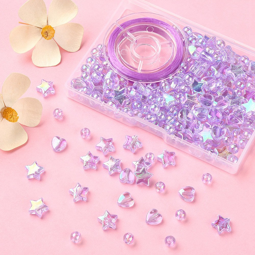 plastic bead box with iridescent light purple transparent beads and spool of purple stretch cord with some loose heart star and round beads spilled out onto pink surface with cream colored flowers in the corner. 