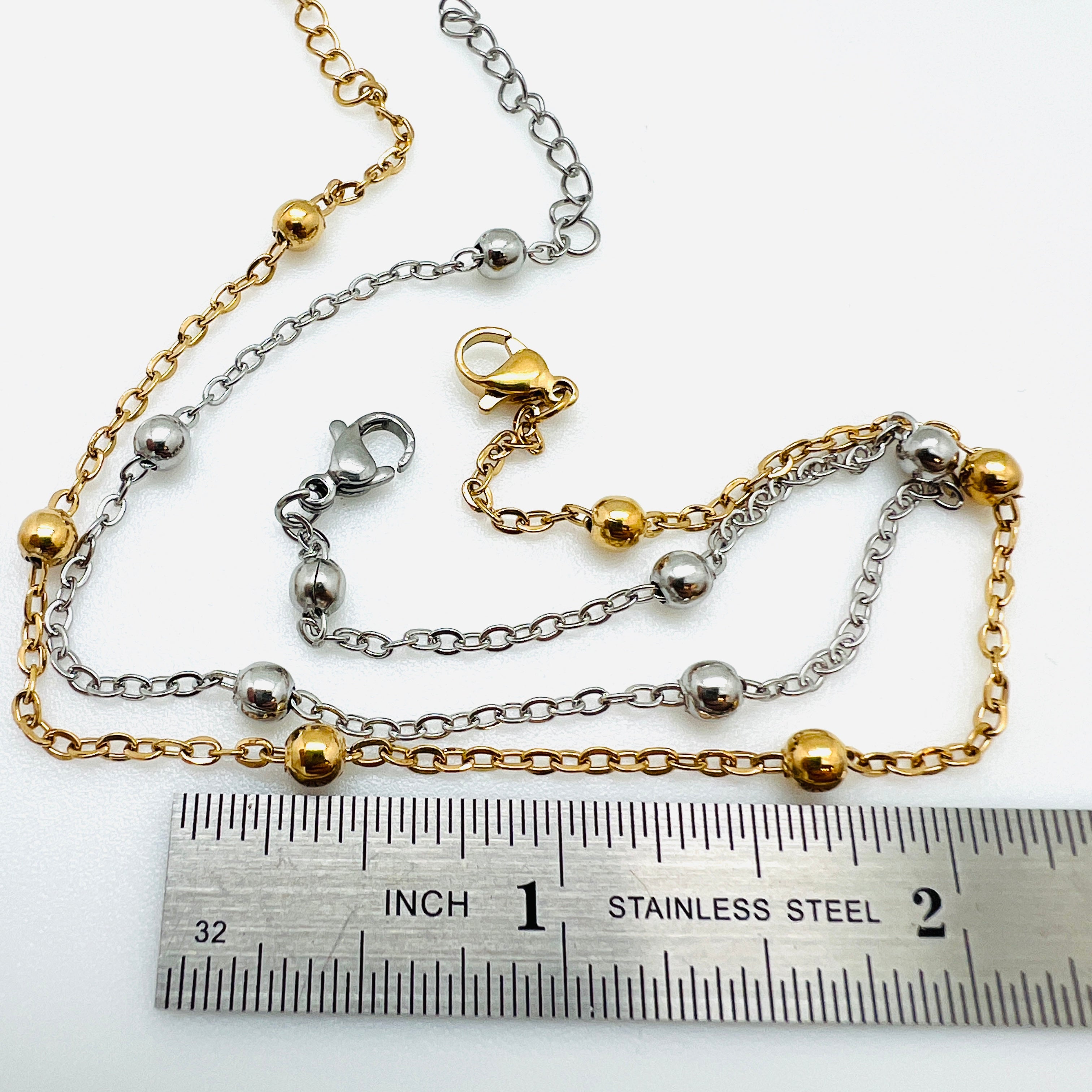 golden and silver tone satellite chain anklets with lobster claw clasp and extender chain displayed above ruler over white background. 