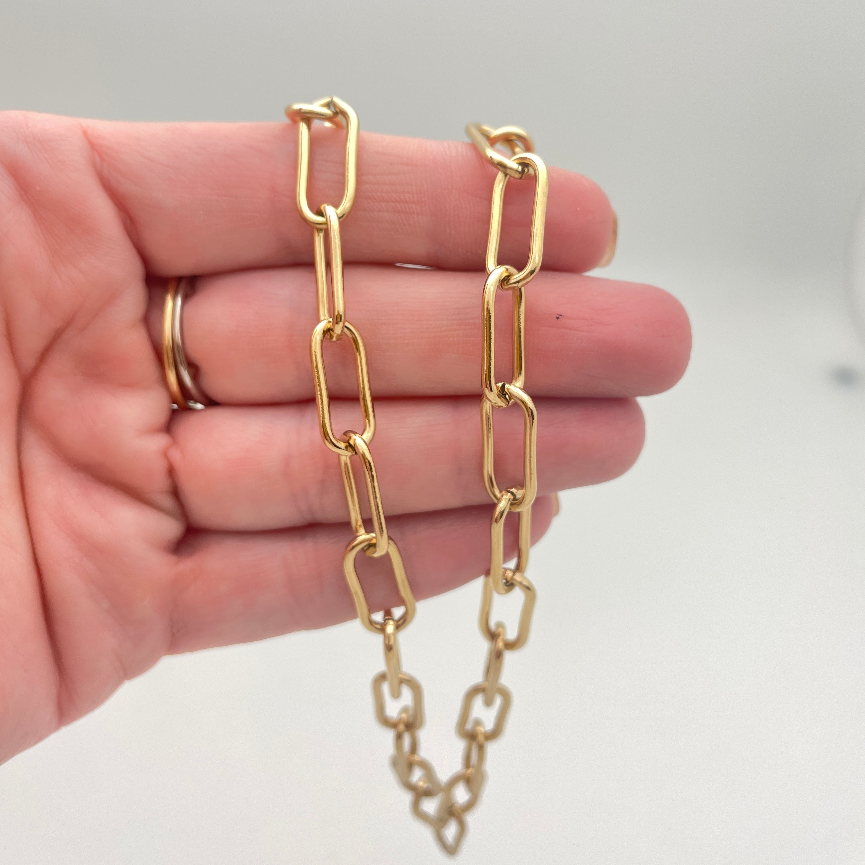 golden large round paperclip chain necklace draped over hand with white background. 