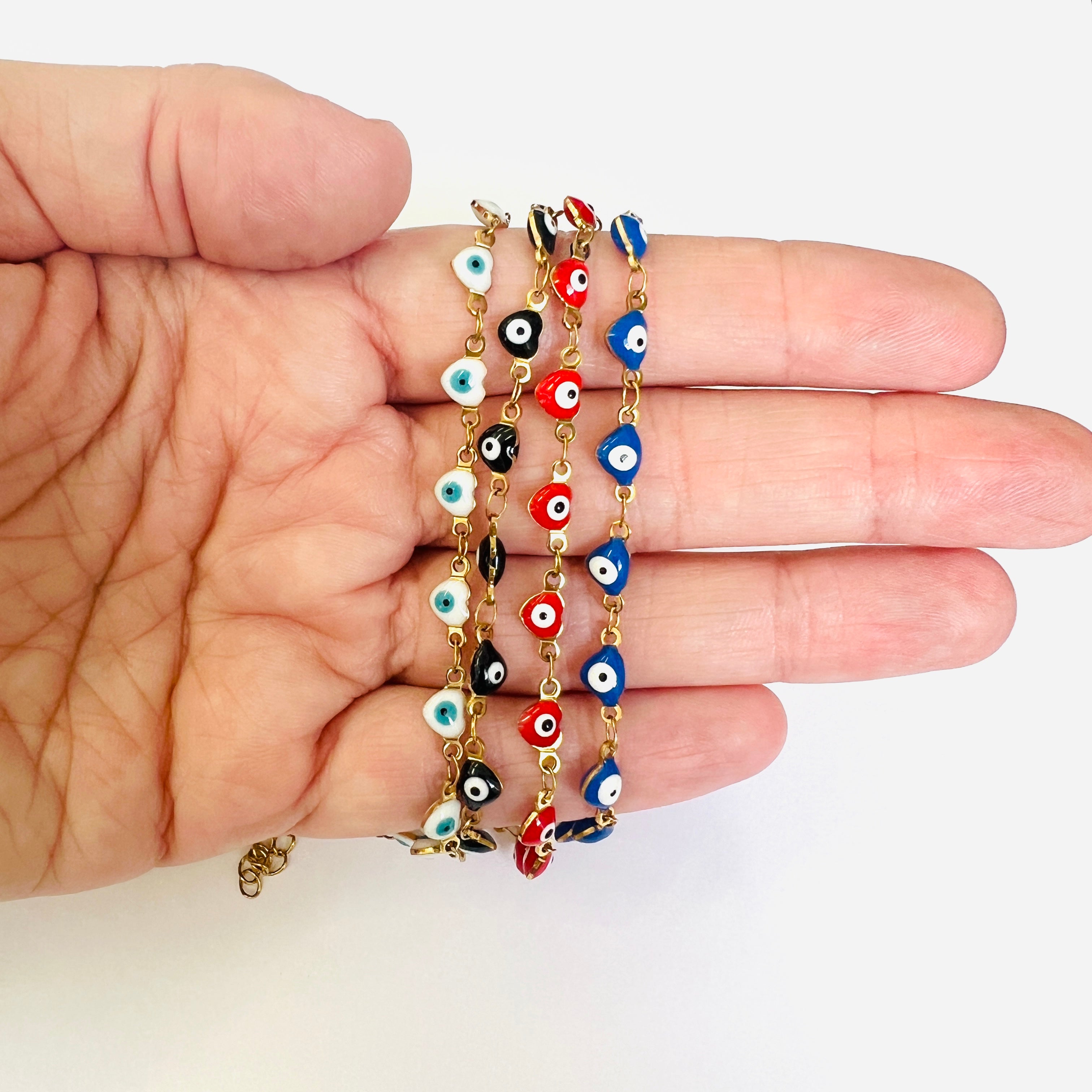 4 golden enamel heart shaped evil eye necklaces showcasing the available color options from left to right  being white, black, red and blue draped over a human hand with white background. 