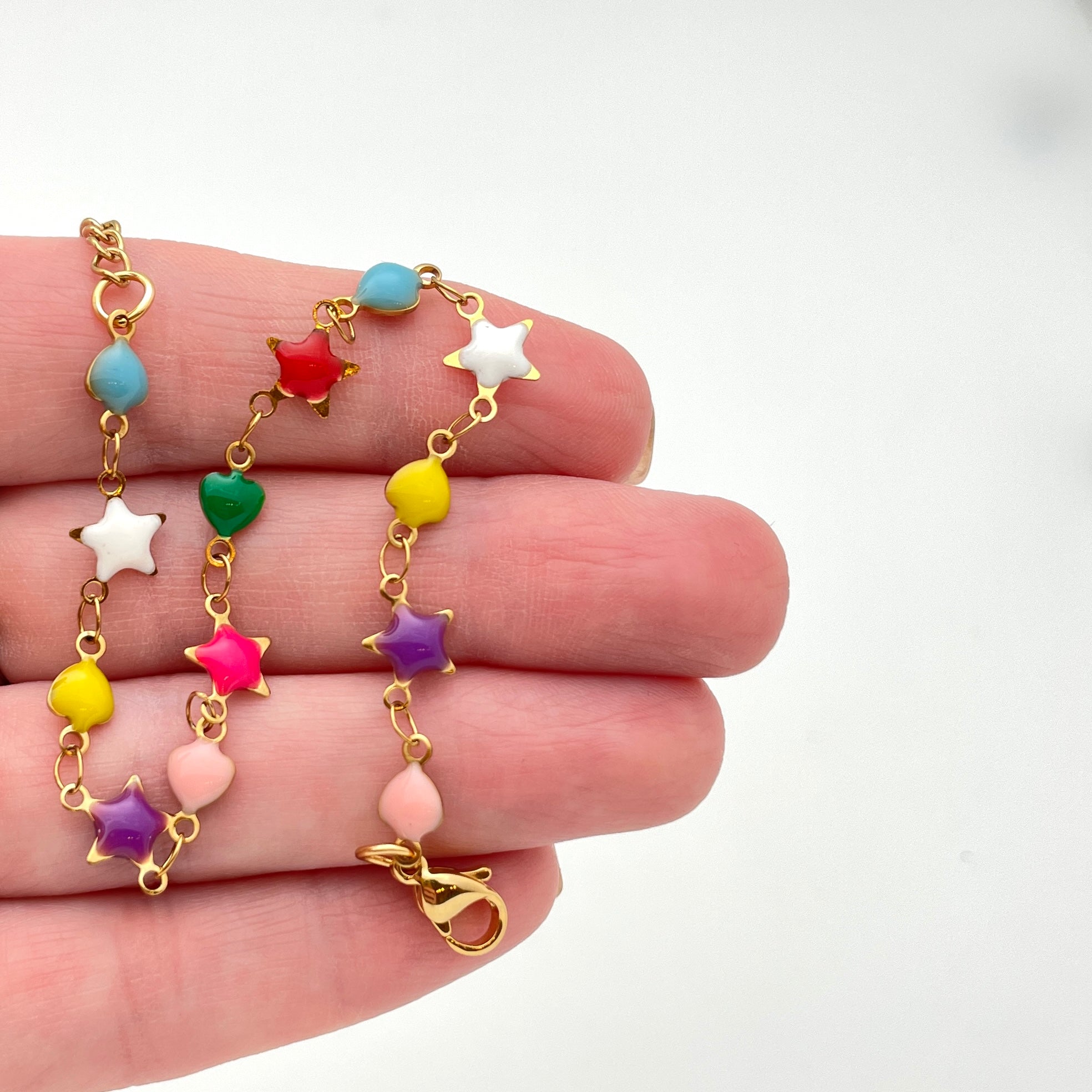 golden multi-colored enamel heart and star link bracelet with lobster claw clasp draped over hand with white background. 