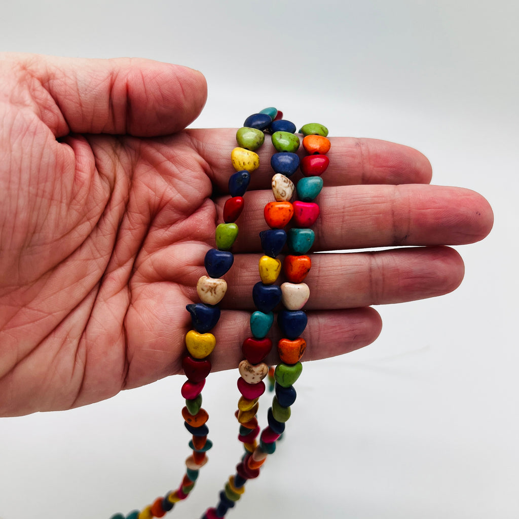 three strands of multicolor dyed magnesite heart stone beads draped over  a hand with a white background. 
