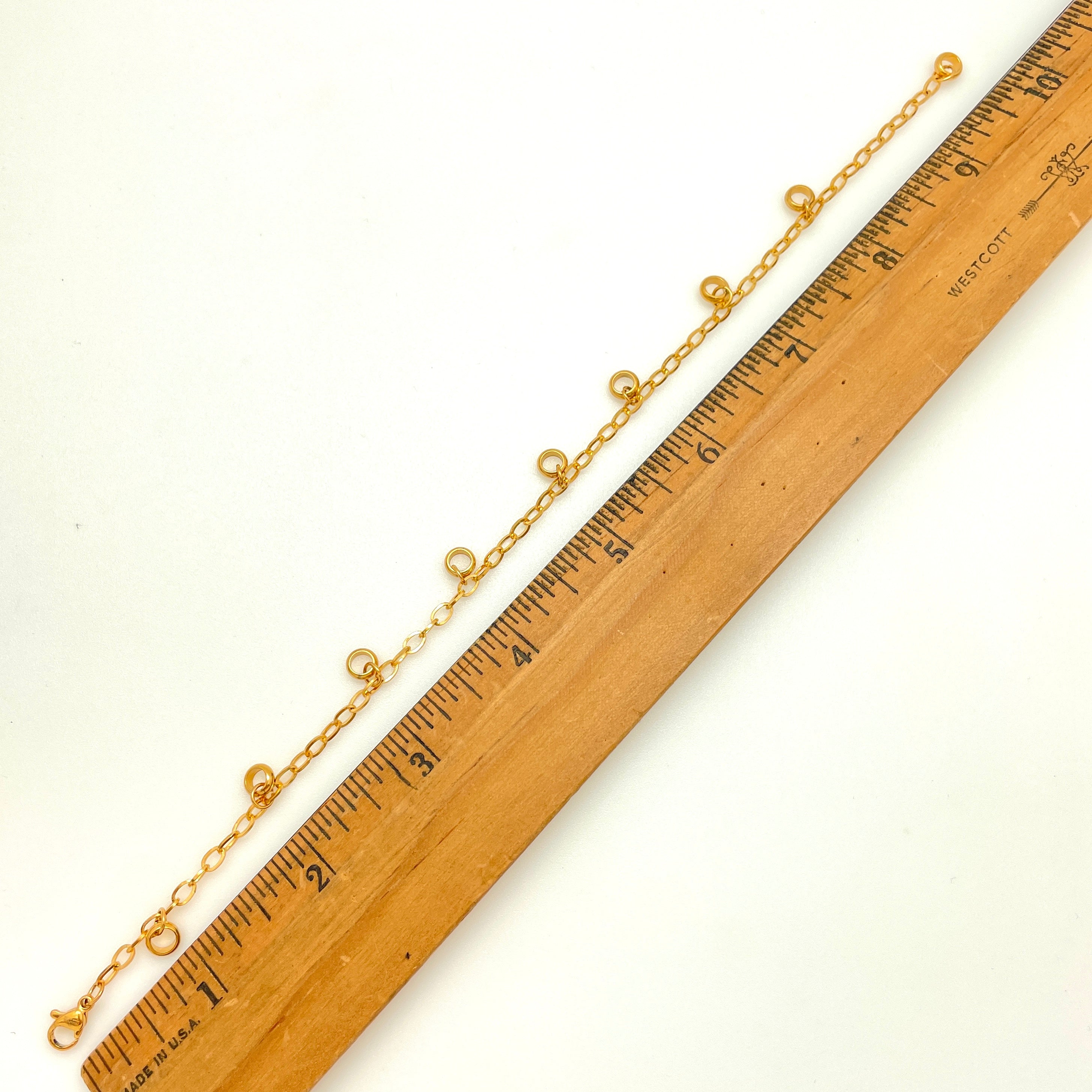 golden add-a-charm bracelet with nine  evenly spaced, dangling, soldered jump rings, a lobster claw clasp and extender chain displayed next to a wooden ruler on white background. 