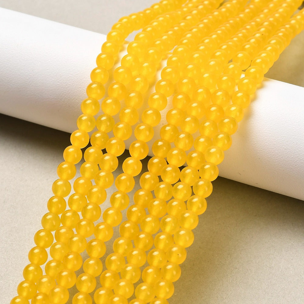 smooth, round, translucent, dyed dark yellow jade bead strands draped over white cylinder form onto off-white  surface. 