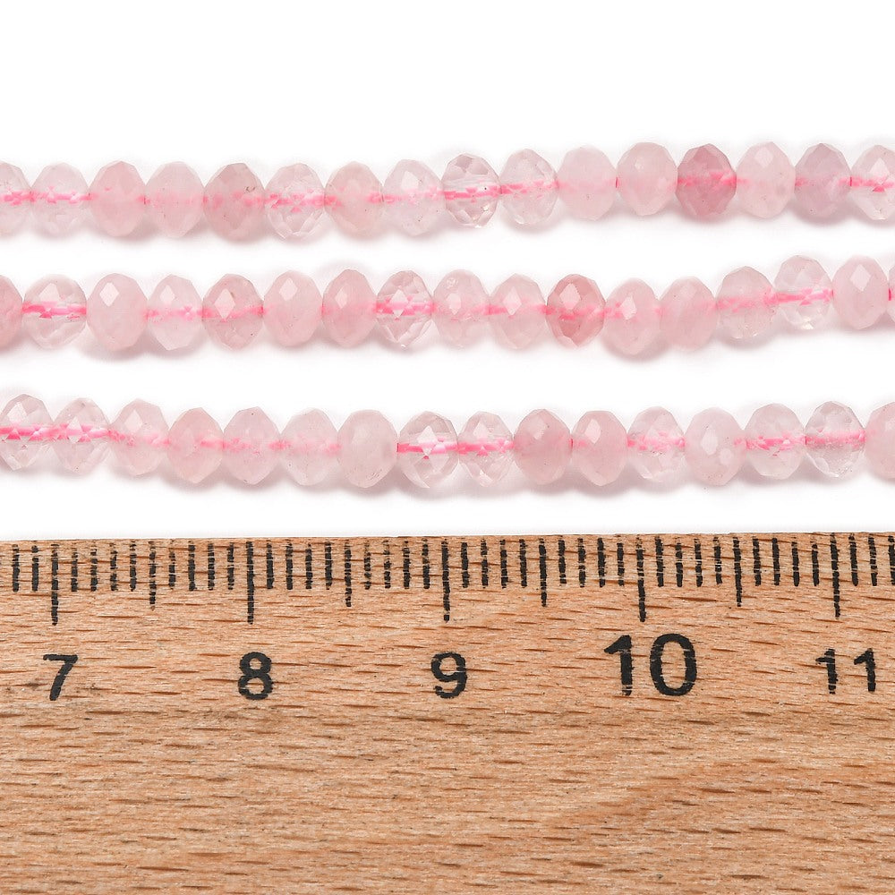 close-up of faceted rondelle rose quartz bead strands above wooden ruler over white background. 