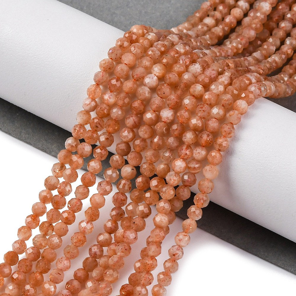 multiple faceted round sunstone bead strands draped over white cylinder from grey onto white surface.