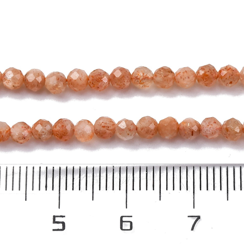 zoomed in sections of two faceted round sunstone bead strands displayed above a ruler with a white background.