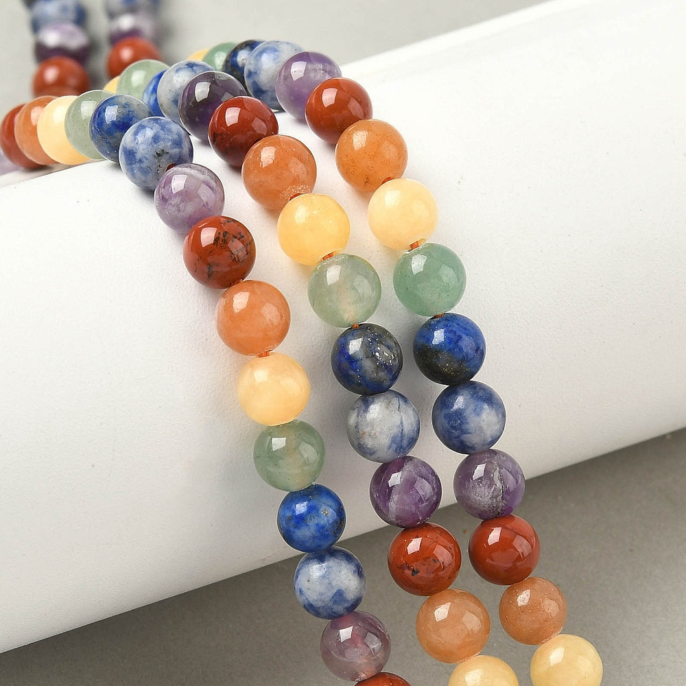 three natural mixed gemstone chakra bead strands draped over white cylinder form onto light grey surface. 