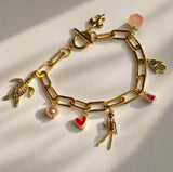 Gold link charm bracelet with assorted charms including a gold turtle, pearl drop, red heart, gold skeleton, hamsa hand with blue stone, peach fruit and gold fleur de lis