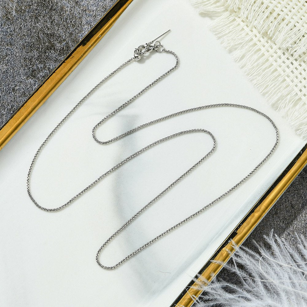 silver tone box chain adjustable slider necklace with lobster claw clasp, slider bead and stopper on gold rimmed tray on grey surface with white tassels and feathers peeking in the corners. 