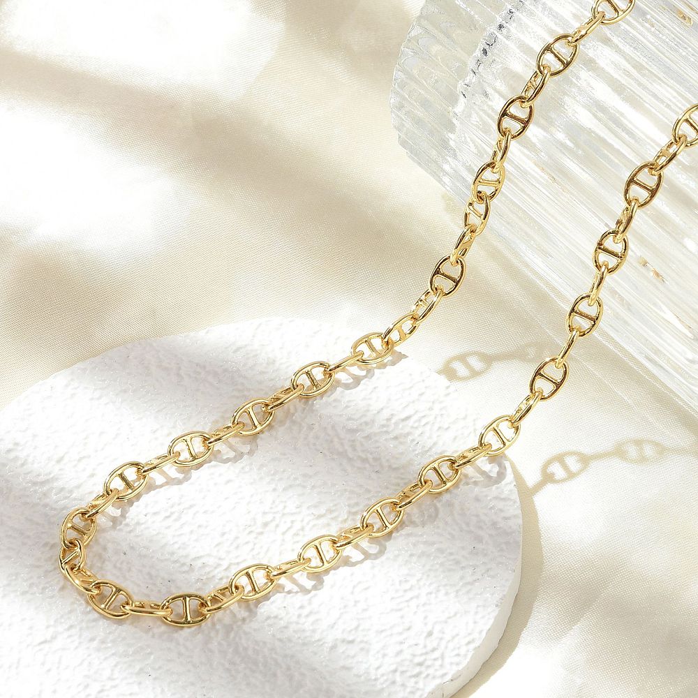 golden oval mariner link chain necklace draped over a crystal glass onto a white textured coasted above an off-white cloth surface. 