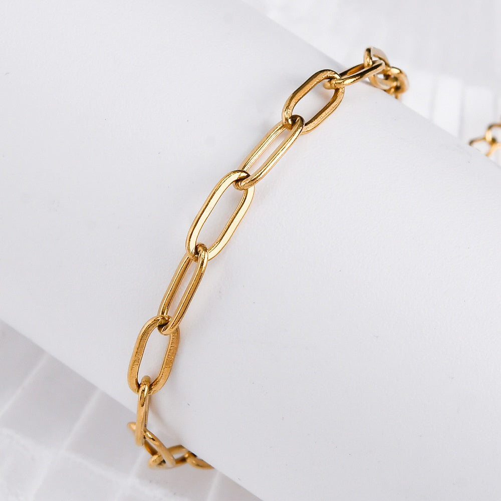 golden paperclip chain bracelet wrapped around white cylinder form above white surface. 