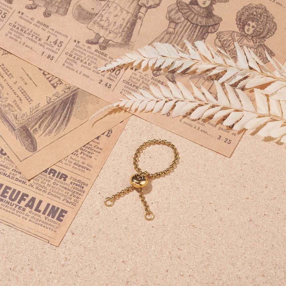 golden adjustable fidget ring with Rolo chain and slider bead and two jump rings over light brown textured surface with old newspaper articles and dried flowers in the top corners. 