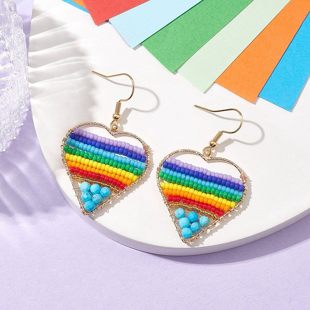 silver tone multicolor dyed magnesite and glass seed bead rainbow heart earrings with ear wires displayed on a white tray with multicolor sheets of paper above them and a light purple surface below. 