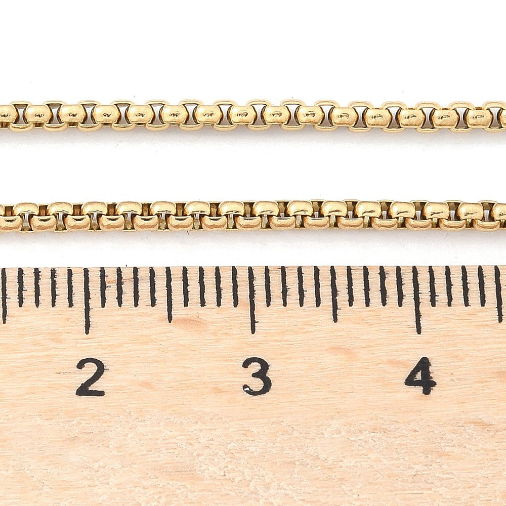 golden Venetian box chain displayed above wooden ruler on white background. 