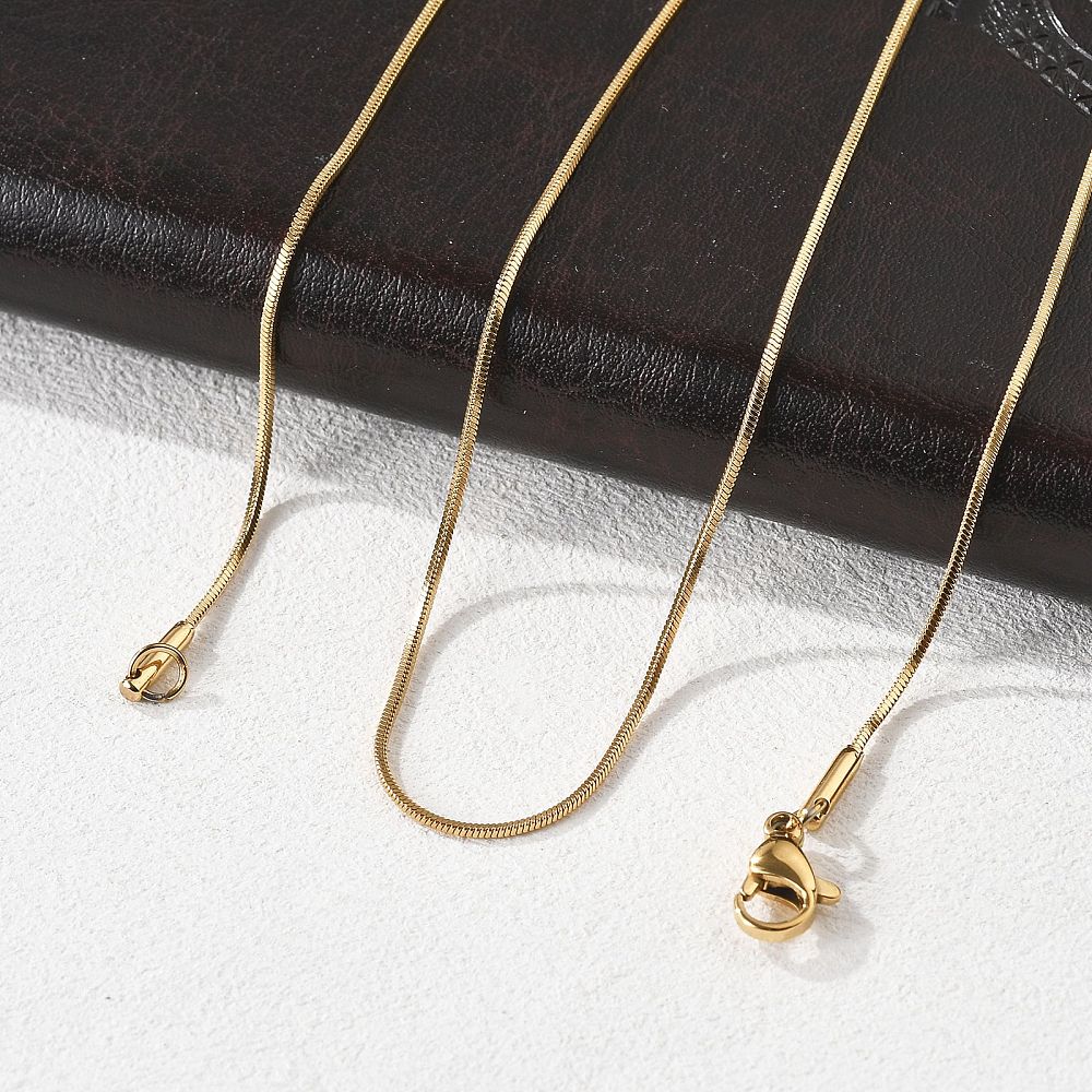 golden square snake chain with lobster claw clasp draped over black leather folder onto a white textured surface. 