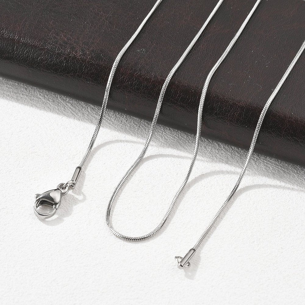 silver tone square snake chain with lobster claw clasp draped over black leather folder onto a white textured surface. 