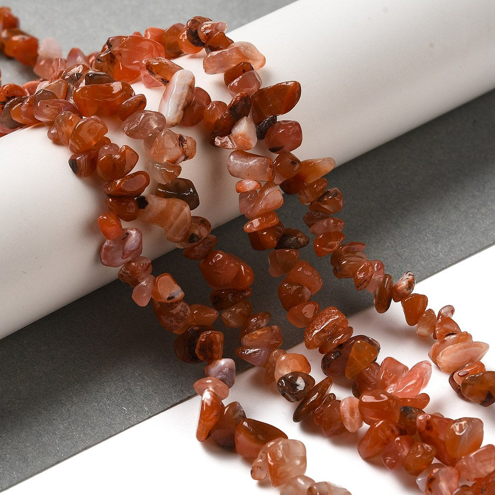 four red agate chip bead strands draped over white cylinder on grey and white surface.