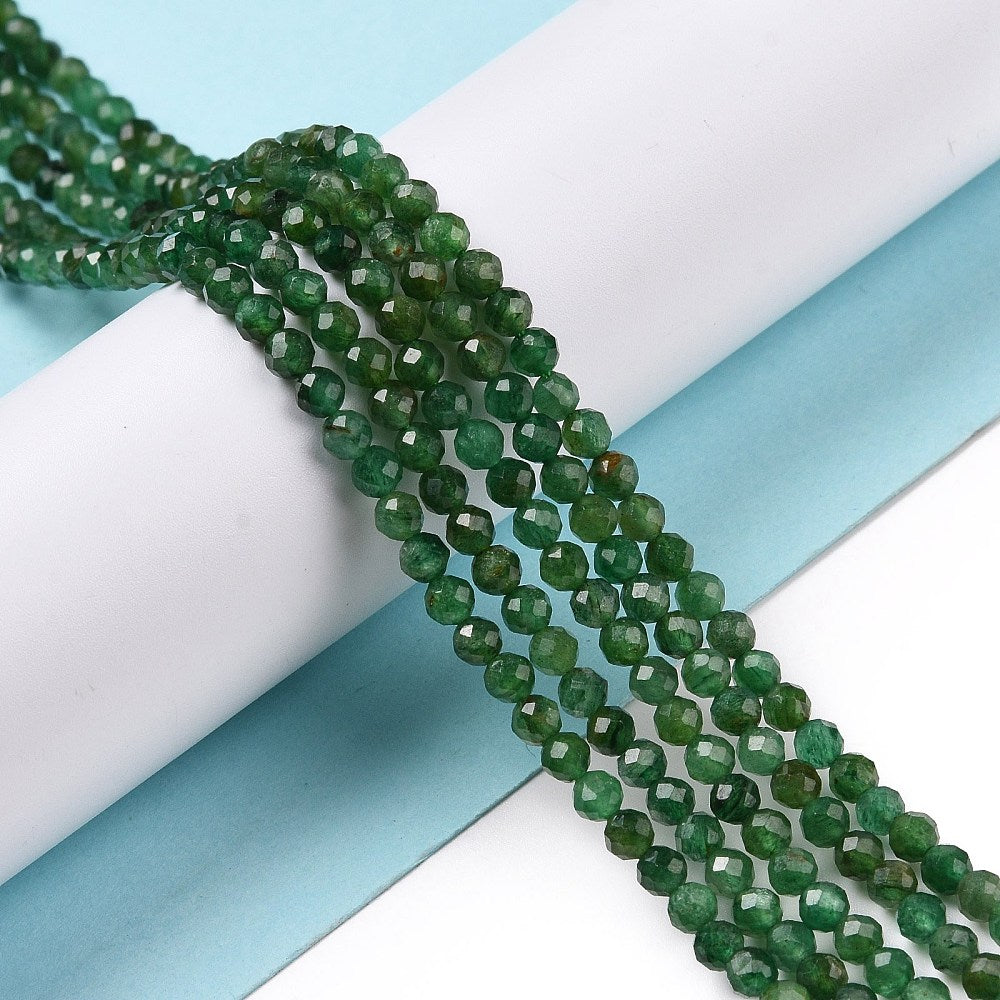 five strands of dark green faceted round Fuchsite beads draped over a white cylinder form above light blue and white surface. 