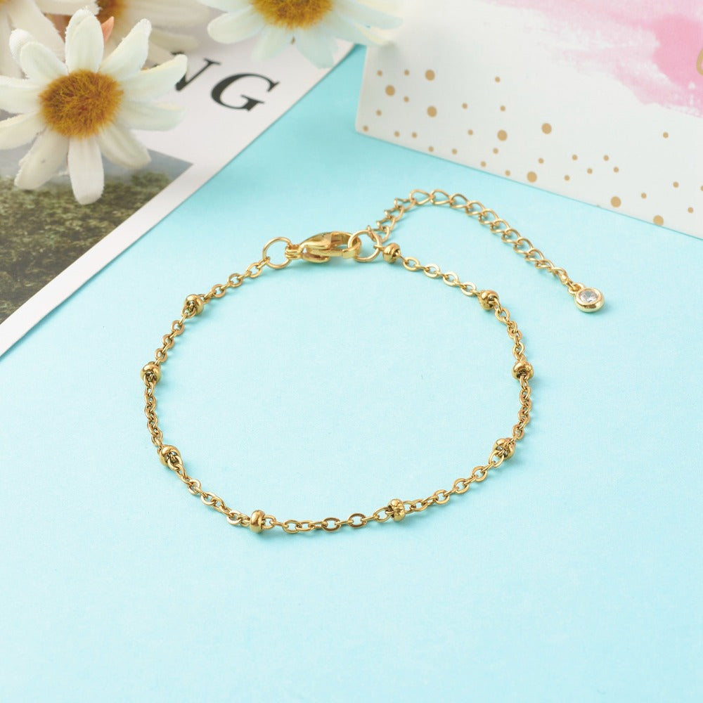 golden satellite chain bracelet with lobster claw clasp and extender chain with bezel set clear stone charm on end on light blue background. 