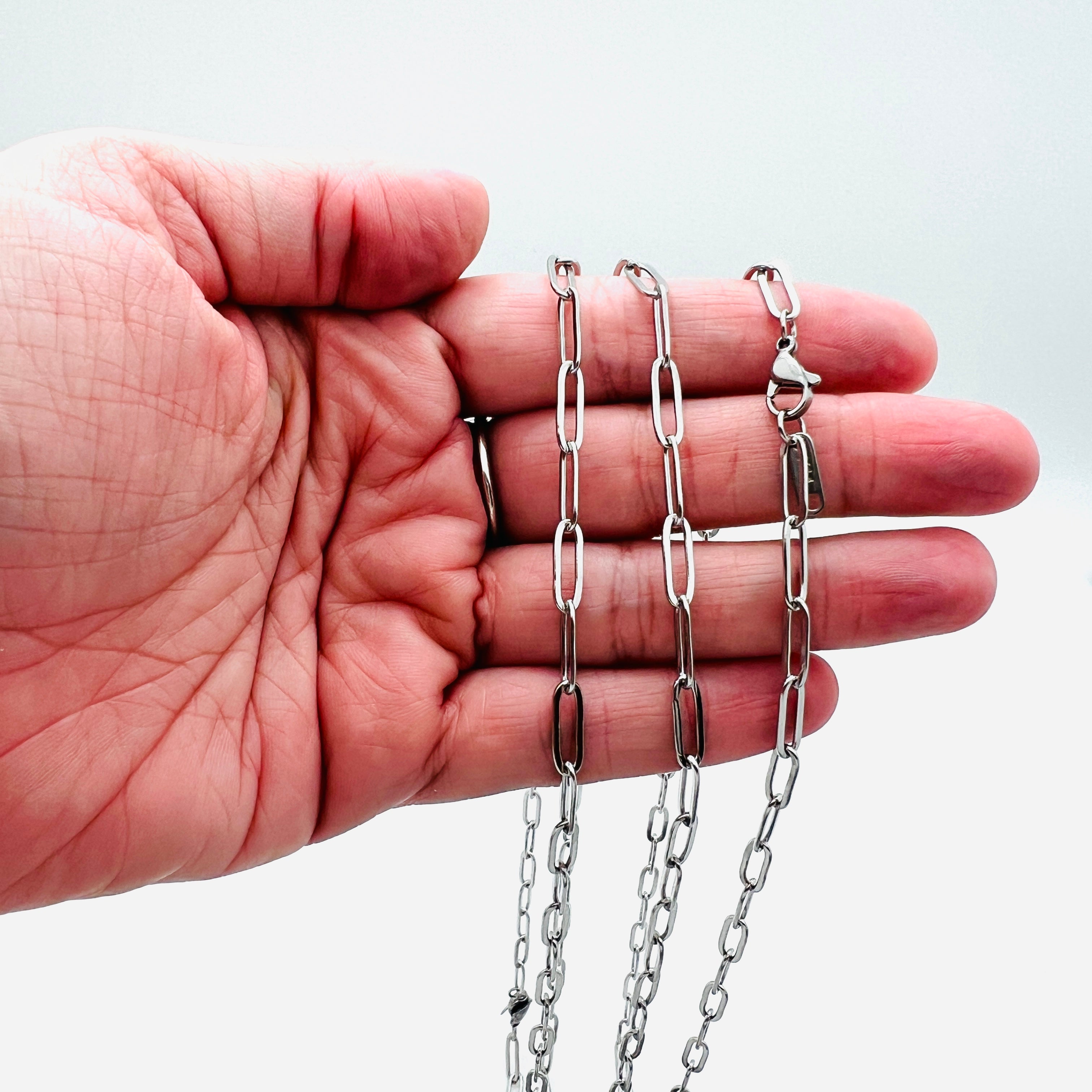 stainless steel medium link paperclip chain necklace draped over a hand with white background. 