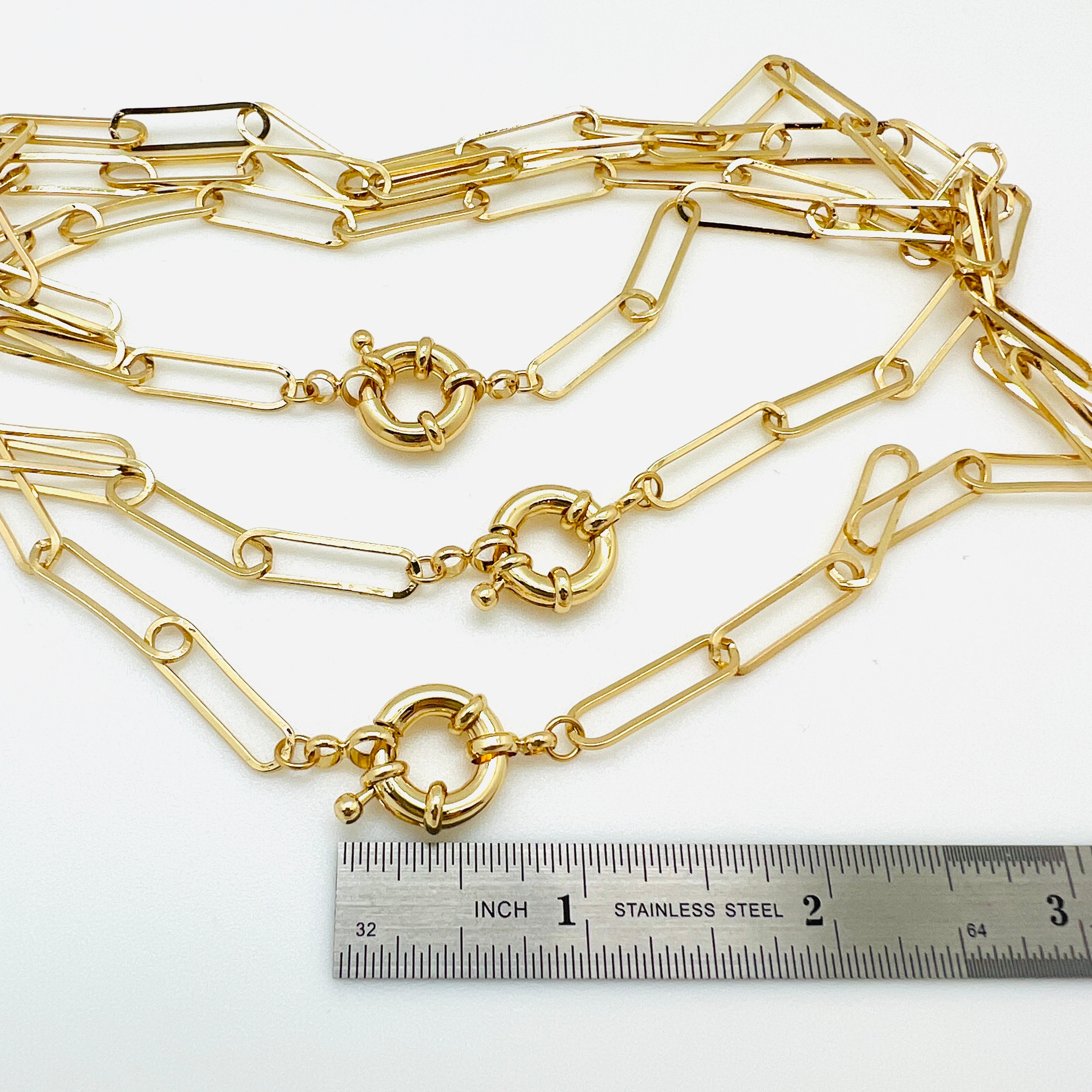three golden paperclip link bracelets with large spring ring clasp displayed above a stainless steel imperial ruler on white background. 