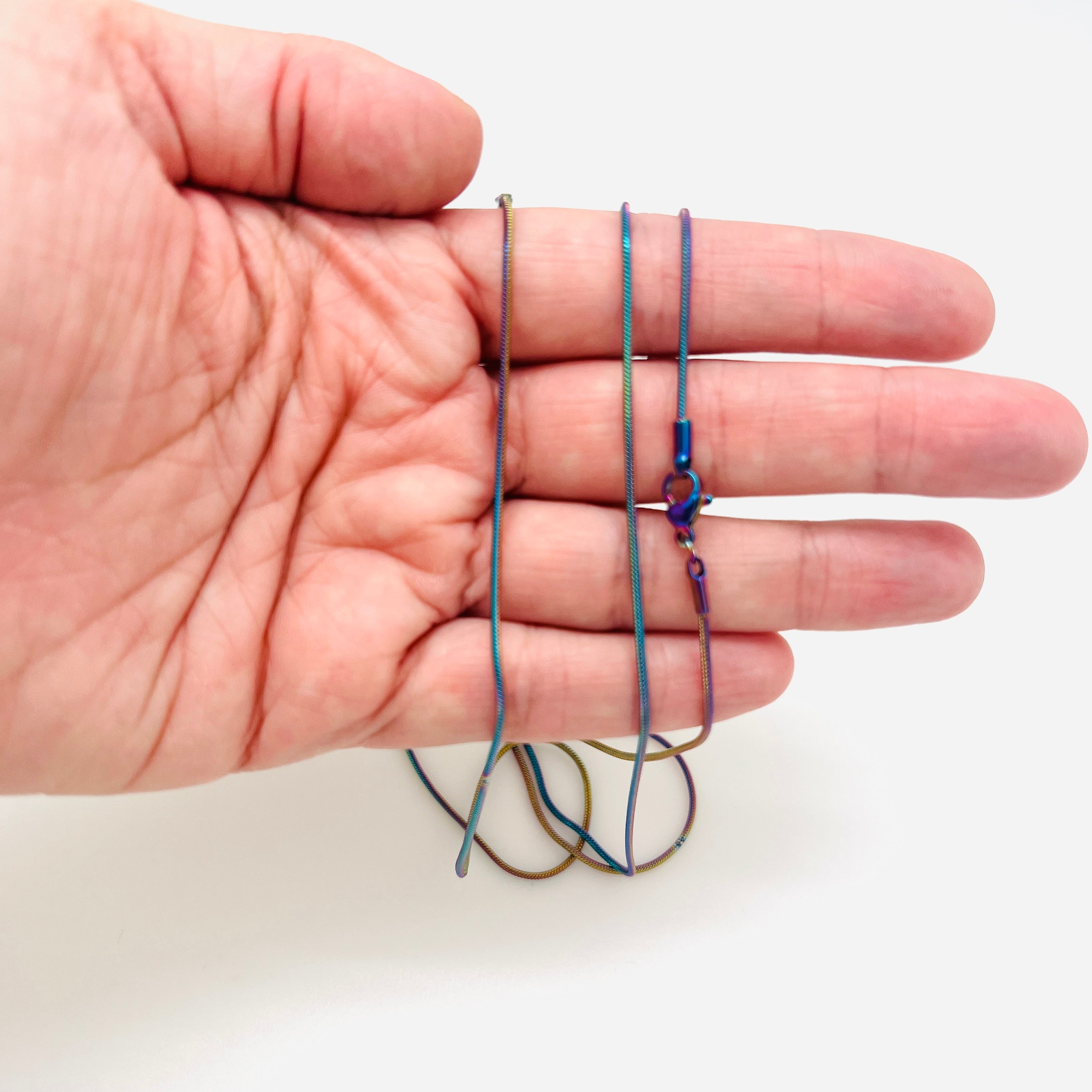 rainbow ion plated snake chain with lobster claw clasp draped over a hand against a white background. 